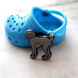 May include: A gray Weimaraner dog shoe charm with black outlines and a red tongue. The charm is attached to a blue Croc shoe.