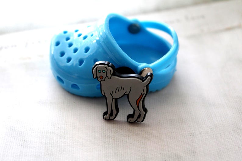 May include: A gray Weimaraner dog shoe charm with black outlines and a red tongue. The charm is attached to a blue Croc shoe.