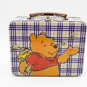 May include: A rectangular metal lunch box featuring a yellow bear wearing a red shirt, surrounded by small bees. The background has a purple and white plaid pattern. The lunch box includes a brown plastic handle and a silver metal latch closure.