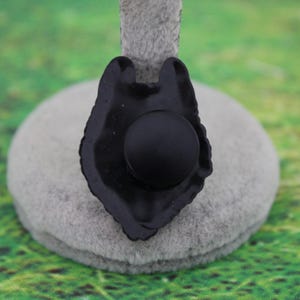 May include: A black, matte-finished pin shaped like a wolf head silhouette. The design features a central circular element and a jagged, stylized outline representing the animal's fur and ears, presented on a neutral gray display stand.