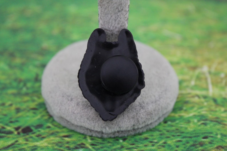 May include: A black, matte-finished pin shaped like a wolf head silhouette. The design features a central circular element and a jagged, stylized outline representing the animal's fur and ears, presented on a neutral gray display stand.