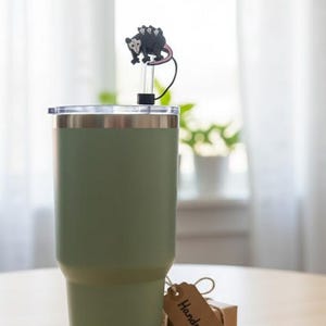 May include: A sage green insulated tumbler with a silver rim and lid. A black and gray opossum straw topper is attached. A small gift box with a "Handmade" tag sits nearby. The tumbler is on a wooden surface.