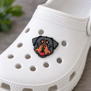 May include: A white foam clog shoe featuring a decorative charm shaped like a Rottweiler dog head. The charm displays a black, tan, and red color palette with a detailed canine face, positioned securely within one of the circular ventilation holes.