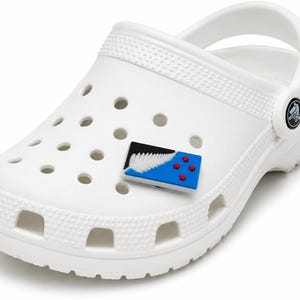 May include: A white foam clog shoe featuring a rectangular charm attached to the upper surface. The charm displays a black and blue flag design with a white silver fern leaf and four red stars, representing the New Zealand national flag.