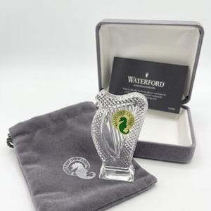 May include: A clear crystal harp figurine featuring a diamond-cut pattern and a green seahorse sticker. It is accompanied by a grey velvet drawstring pouch with a seahorse logo and a grey hinged presentation box containing a Waterford brand card.