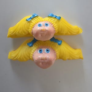 May include: Two plastic hair clips featuring molded doll faces with bright yellow hair, blue bows, and blue eyes. Each clip has a peach-toned face with a small nose and mouth, designed to hold hair in place with the attached yellow plastic bar.