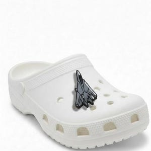 May include: White clogs with a gray and black airplane charm. The clogs have multiple ventilation holes and a textured surface. The airplane charm is a detailed miniature of a jet.