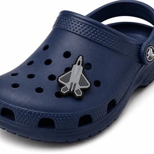 May include: A navy blue clog with a textured surface and ventilation holes. A gray and black fighter jet charm is attached to the top of the shoe. The clog features a pivoting heel strap with a circular logo button on the side of the strap.