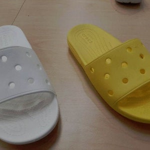May include: Two pairs of Crocs slides, one in white and one in yellow. Both pairs have a textured, perforated design and the brand name "Crocs" is visible on the yellow pair.