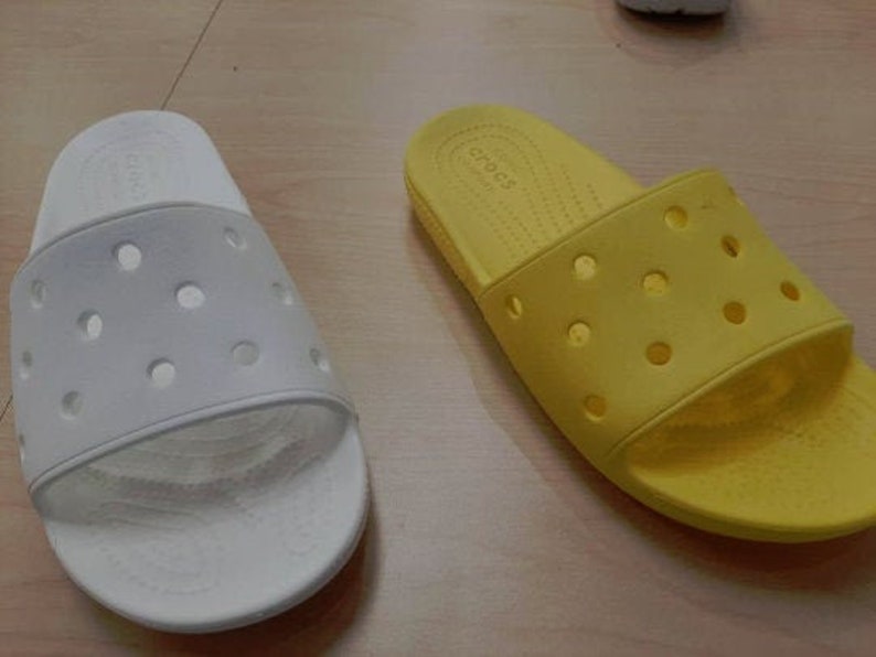 May include: Two pairs of Crocs slides, one in white and one in yellow. Both pairs have a textured, perforated design and the brand name "Crocs" is visible on the yellow pair.