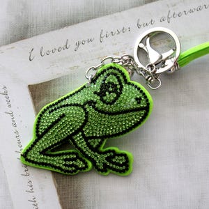 May include: A green frog keychain with rhinestones, attached to a silver key ring with a green cord.