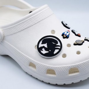 May include: A white foam clog shoe featuring a collection of space-themed charms inserted into the ventilation holes. The charms include a black circular alien silhouette, a colorful rocket ship, a grey planet with rings, and a small star.