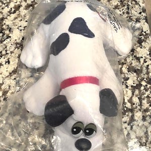 May include: A white plush dog toy with black spots, a red collar, and black ears, enclosed in a clear plastic bag. The bag features printed text that reads CAUTION and includes safety warnings regarding plastic bags and suffocation hazards.