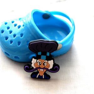 May include: A light blue clog shoe with a cartoon chef-themed shoe charm. The charm features a chef with a white mustache, a purple hat, and a blue and black uniform. The shoe has multiple holes.