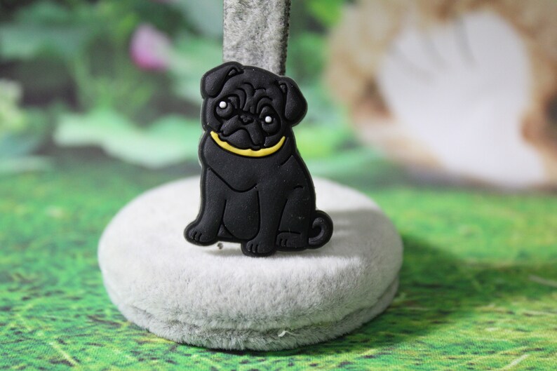 May include: Black rubber pug charm with a yellow collar. The charm is sitting on a gray surface.