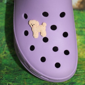 May include: A purple clog with a small, beige dog charm attached to the front. The clog has a textured surface and black holes around the sides.
