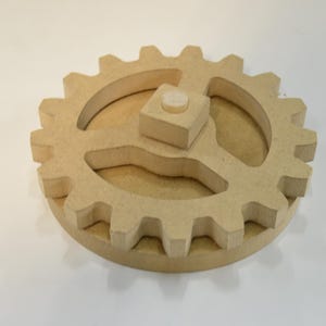 May include: A wooden gear with interlocking teeth and a central square block with a circular peg. The gear is made of light brown wood and is designed for mechanical or educational purposes.