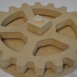 May include: A wooden gear with interlocking teeth and a central square block with a small peg. The gear is made of light brown wood and has a three-dimensional design. The product is likely a puzzle or a decorative item.