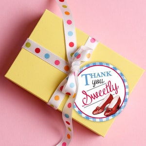 Może przedstawiać: Żółte pudełko prezentowe z białą wstążką w kolorowe kropki. Do wstążki przymocowana jest okrągła etykieta w niebiesko-białe kratki. Etykieta zawiera napis "Thank you Sweetly" i obrazek rubinowych pantofelków.