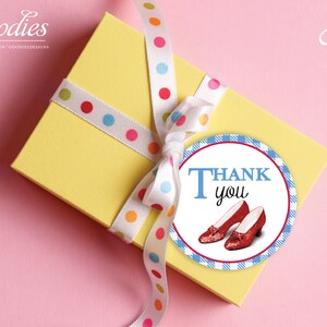 Może przedstawiać: Żółte pudełko prezentowe z białą wstążką w kolorowe kropki. Pudełko ma okrągłą etykietę z podziękowaniem na niebiesko-białym tle w kratkę. Etykieta zawiera napis "THANK you" niebieskimi literami i obrazek rubinowych pantofelków.