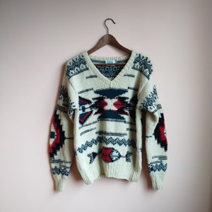 May include: A cream-coloured knitted sweater with a V-neck and a geometric pattern in red, blue, and black. The sweater has long sleeves and a relaxed fit.