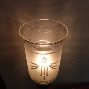 May include: A frosted glass lampshade with a decorative design of a stylised bird with wings spread and a hanging pendant. The lampshade is lit from within, casting a warm glow.