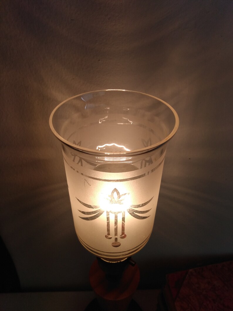 May include: A frosted glass lampshade with a decorative design of a stylised bird with wings spread and a hanging pendant. The lampshade is lit from within, casting a warm glow.