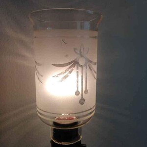 May include: A frosted glass lampshade with a white etched design of stylised leaves and a dangling chain of circles. The lampshade is lit from within, casting a warm glow.