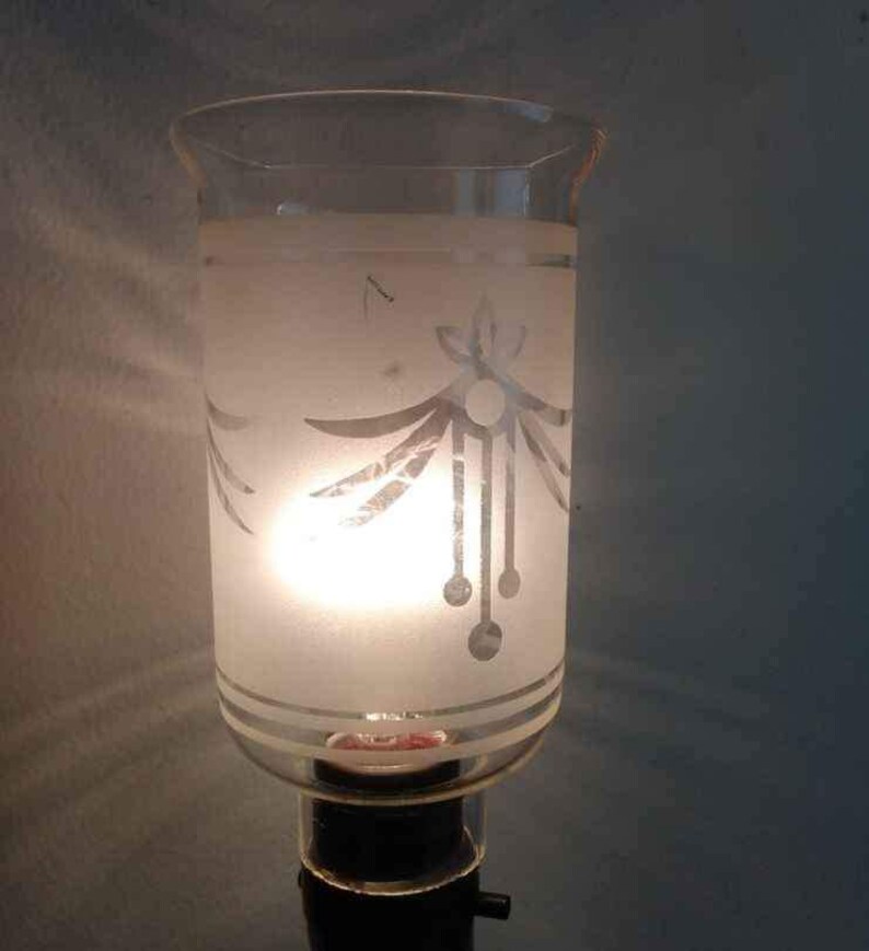 May include: A frosted glass lampshade with a white etched design of stylised leaves and a dangling chain of circles. The lampshade is lit from within, casting a warm glow.
