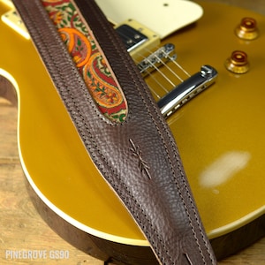 May include: A brown leather guitar strap with a paisley patterned fabric insert. The strap is stitched with white thread and has a small logo on the leather.