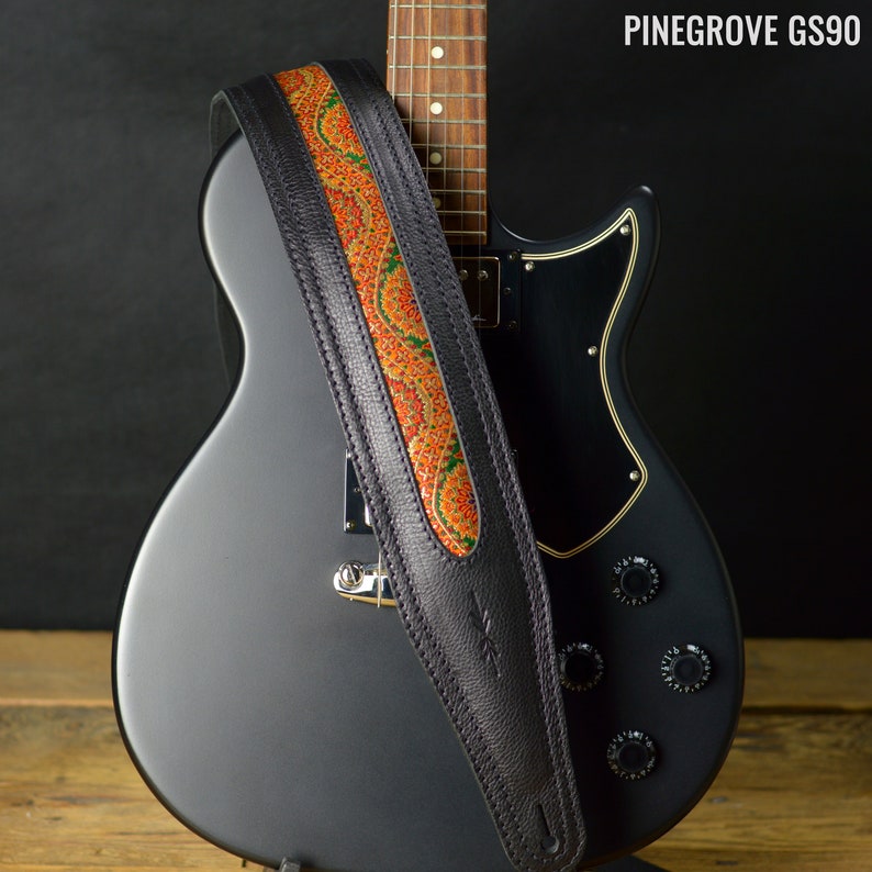 May include: A black electric guitar with a black leather guitar strap with an orange and green paisley design. The strap is labeled "Pinegrove GS90".