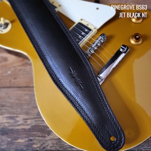May include: Black leather guitar strap with a stitched edge and a small logo. The strap is on a gold electric guitar.