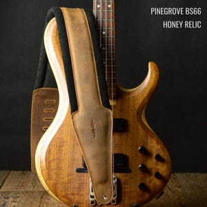 May include: A honey-colored bass guitar with a brown leather strap. The strap is black on the underside and has a decorative stitched design. The text "PINEGROVE BS66 HONEY RELIC" is visible in the upper right corner.