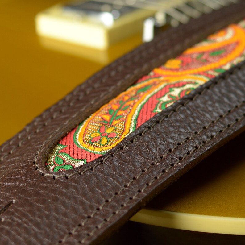 May include: Close-up of a brown leather guitar strap with a paisley patterned fabric insert. The strap is stitched with brown thread.