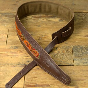 May include: A brown leather guitar strap with a colourful paisley pattern. The strap has a brown leather adjustment strap and a brown leather end strap.