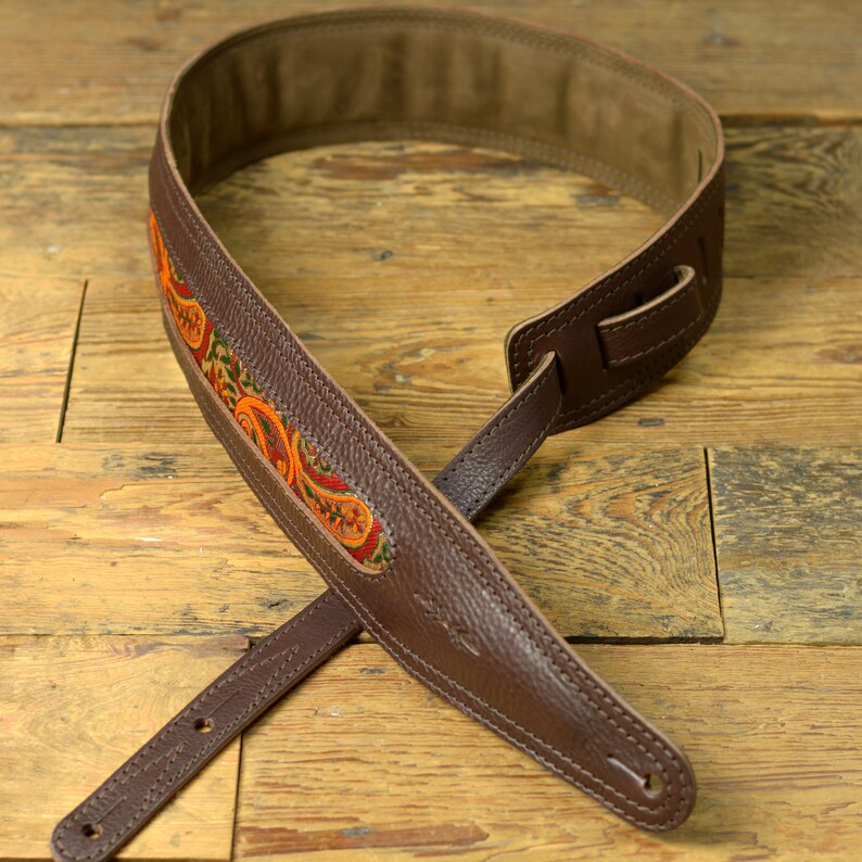 May include: A brown leather guitar strap with a colourful paisley pattern. The strap has a brown leather adjustment strap and a brown leather end strap.