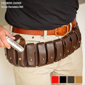 Pode incluir: Um cinto de couro marrom para gaita com oito bolsas individuais para guardar gaitas. O cinto é usado na cintura e tem uma fivela de couro marrom. O texto "PINEGROVE LEATHER Deluxe Harmonica Belt" é visível na parte superior da imagem.