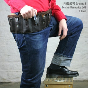 Op de afbeelding: Een bruine leren harmonica riem met acht vakken voor harmonicas. De riem wordt gedragen door iemand die een rood shirt en een blauwe jeans draagt. PINEGROVE Straight-8 Leather Harmonica Belt & Case staat in witte tekst op een zwarte achtergrond.