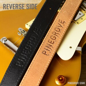 May include: Two guitar straps, one black leather and one brown leather, with the text "Pinegrove Made in England" embossed on the brown strap. The straps are lying on top of a gold guitar.