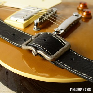 May include: Close-up of a black leather guitar strap with a silver buckle. The strap is attached to a gold-colored electric guitar.