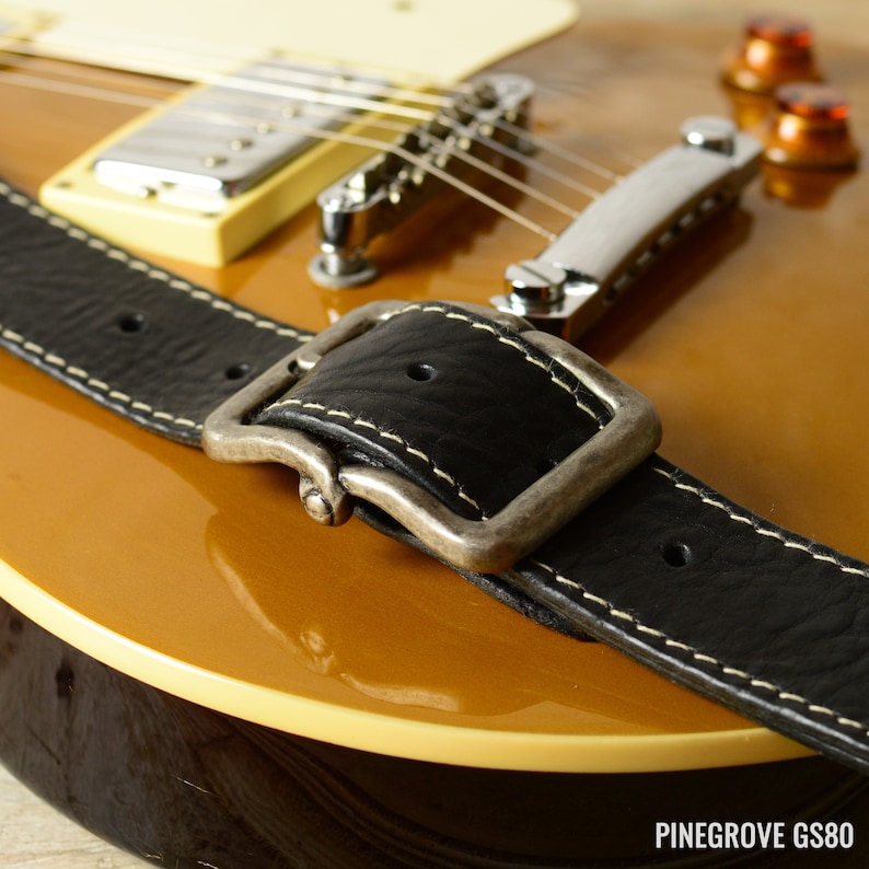 May include: Close-up of a black leather guitar strap with a silver buckle. The strap is attached to a gold-colored electric guitar.