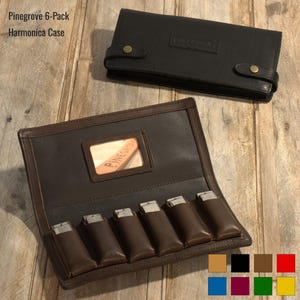 May include: A brown leather harmonica case with a clear window displaying the Pinegrove logo. The case has six slots for harmonicas and a flap with a snap closure. The case is shown on a wooden surface.