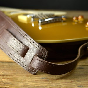 May include: Close-up of a brown leather guitar strap with stitching detail. The strap is resting on a wooden surface.