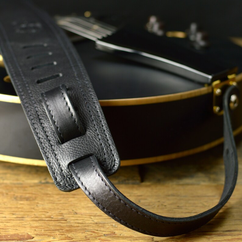 May include: Black leather guitar strap with a textured surface and stitching detail. The strap is attached to a black guitar with gold hardware.