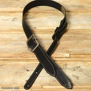 May include: Black leather guitar strap with a silver buckle and white stitching. PINEGROVE GS80 is printed on the strap.