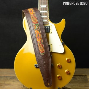 May include: A brown leather guitar strap with a colourful paisley pattern. The strap is attached to a gold electric guitar.