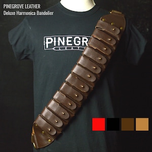 Può includere: Un bandolere in pelle marrone per armonica con dettagli in ottone e più tasche. Il bandolere è indossato su una maglietta nera con la scritta "Pinegrove Leather" stampata sul davanti.