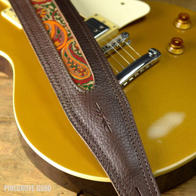 May include: A brown leather guitar strap with a paisley patterned fabric insert. The strap is stitched with white thread and has a small logo on the leather.