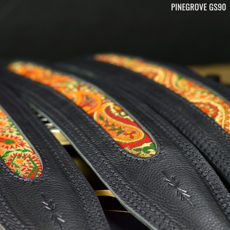 May include: Three black leather guitar straps with colorful paisley patterned fabric inserts. The straps are stitched with white thread and have a small logo on the bottom.