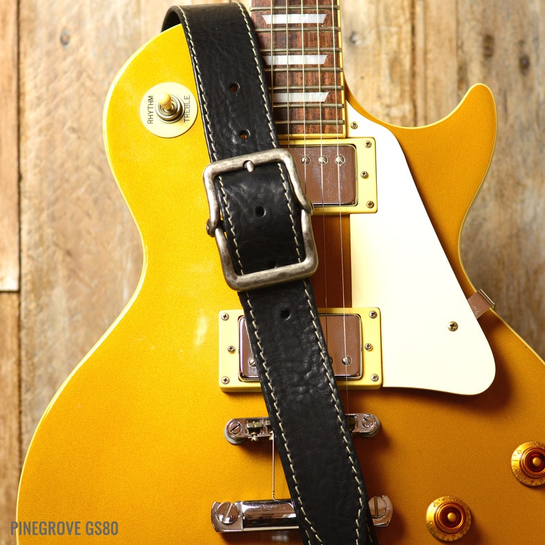 May include: A black leather guitar strap with a stitched edge and a silver buckle. The strap is on a yellow electric guitar with a white pickguard. The guitar has a "PINEGROVE GS80" label on the lower left side.